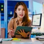 A professional woman sitting at a desk, holding a tablet and discussing insights, representing the idea of transforming experience into valuable coaching content.