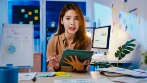 A professional woman sitting at a desk, holding a tablet and discussing insights, representing the idea of transforming experience into valuable coaching content.