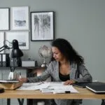 Focused businesswoman working on documents at her desk, surrounded by paperwork and modern office decor.